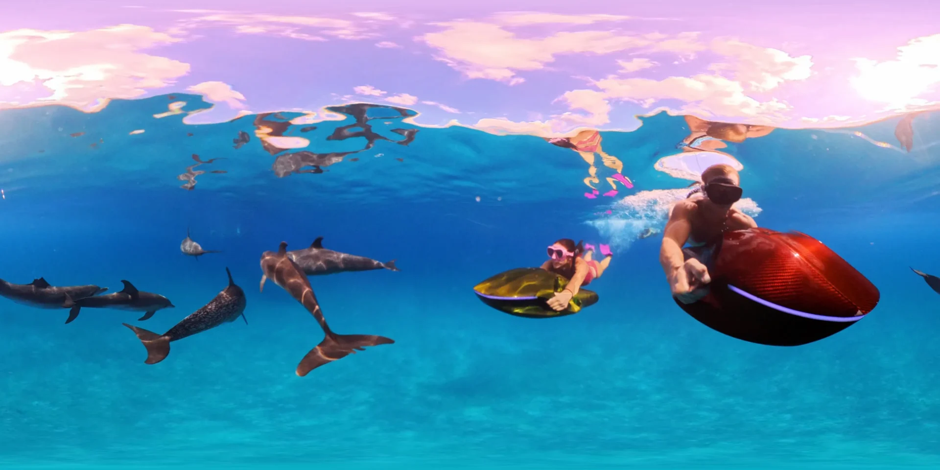 Two snorkelers glide underwater beside a group of dolphins, each holding a sleek electric sea scooter—one red and one gold—beneath a calm, pink-tinted ocean surface with sunlight filtering through the water.