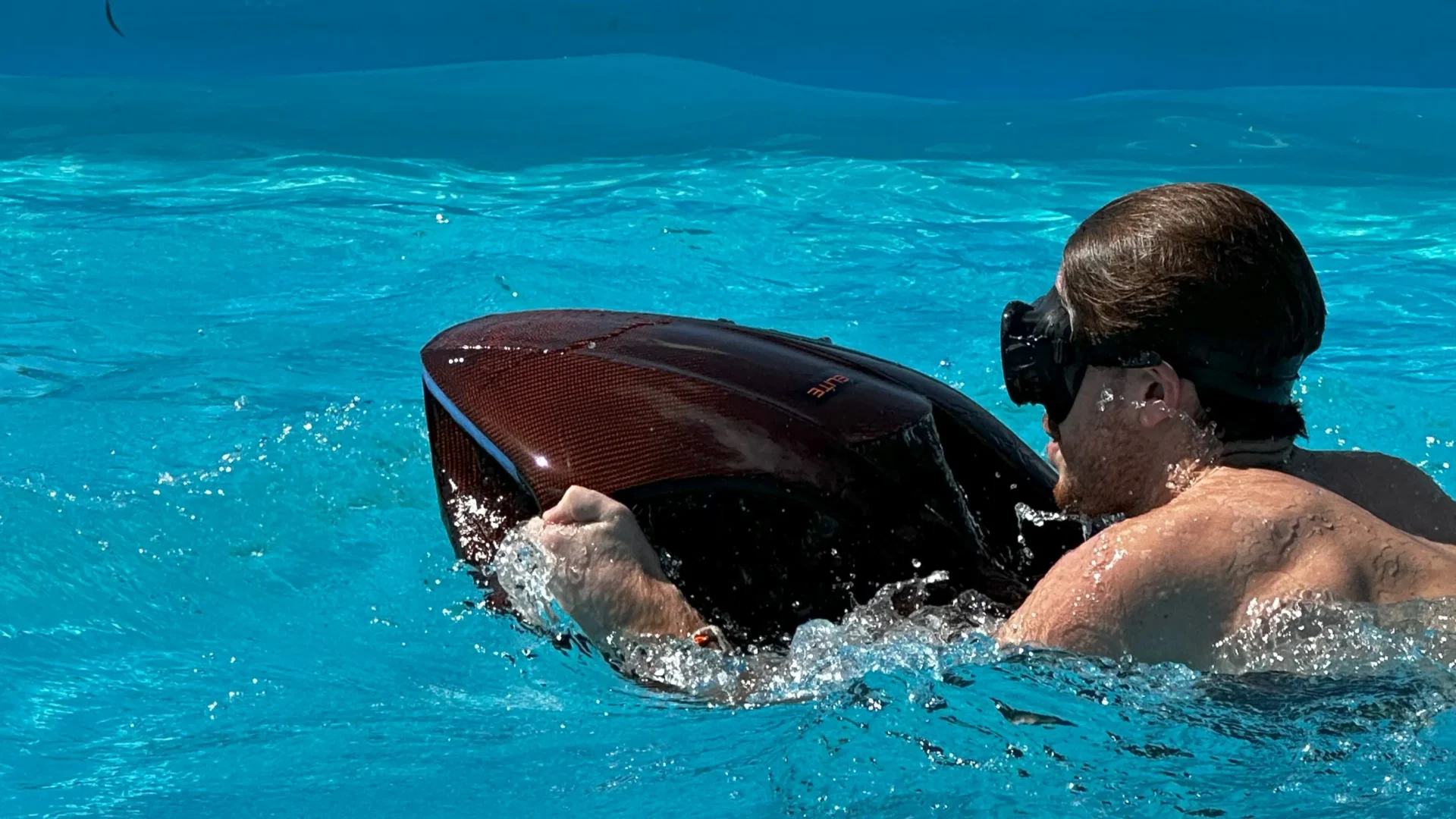 A man wearing swim goggles tests a red carbon fiber electric sea scooter in a bright blue pool, holding onto the handles as water splashes around the device.
